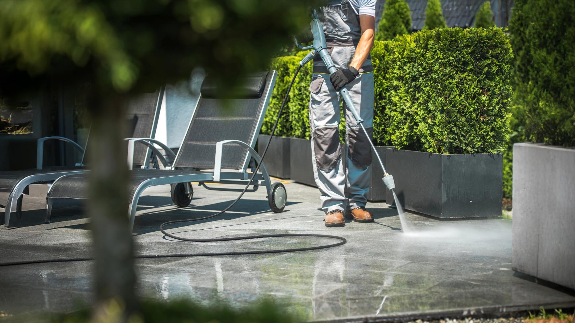 A person in gray overalls uses a pressure washer to clean a stone patio, with a lounge chair nearby and neatly trimmed hedges in the background.